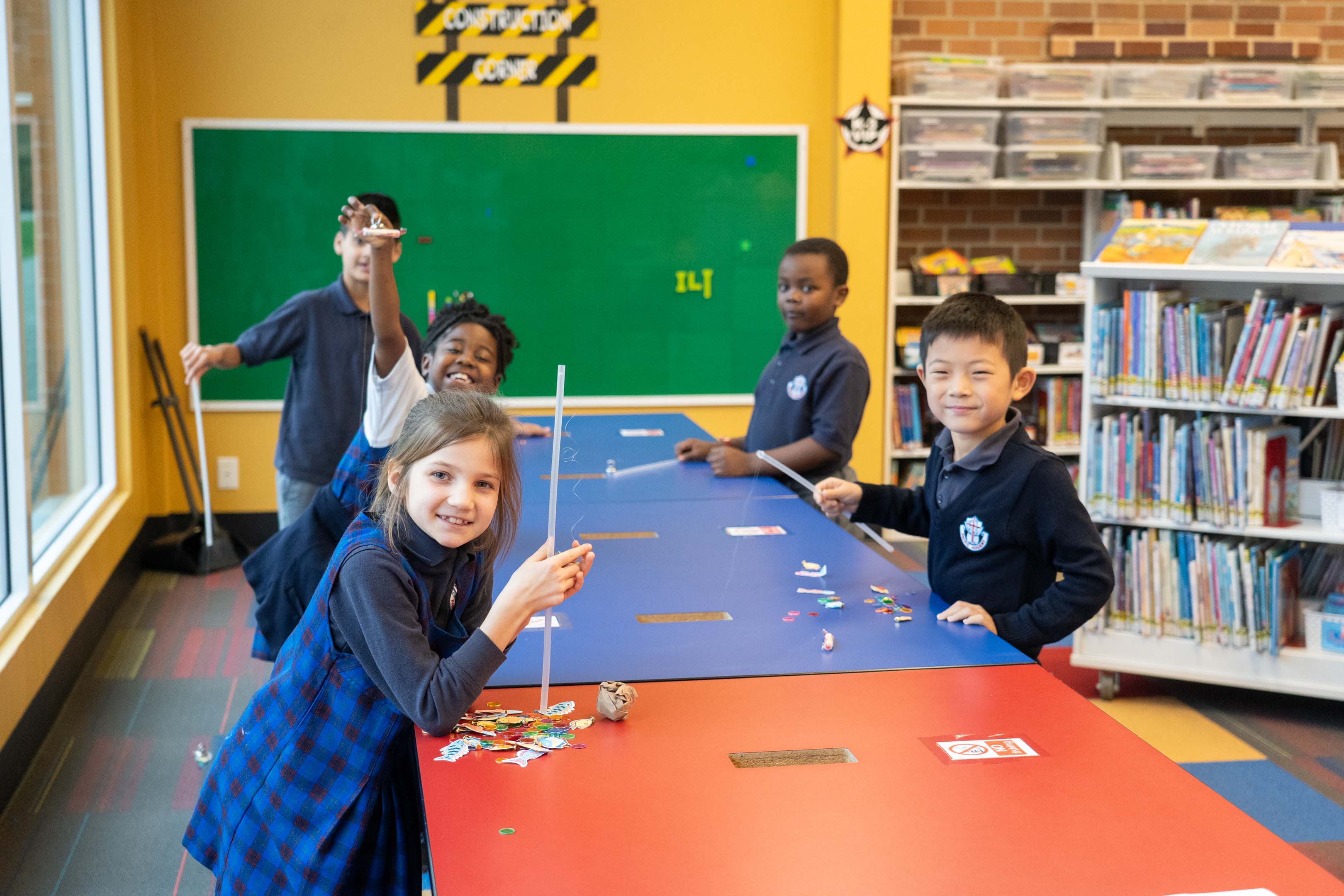 Five diverse children collaborate on a colorful classroom table activity, smiling and holding craft pieces, with a chalkboard and bookshelves behind them.