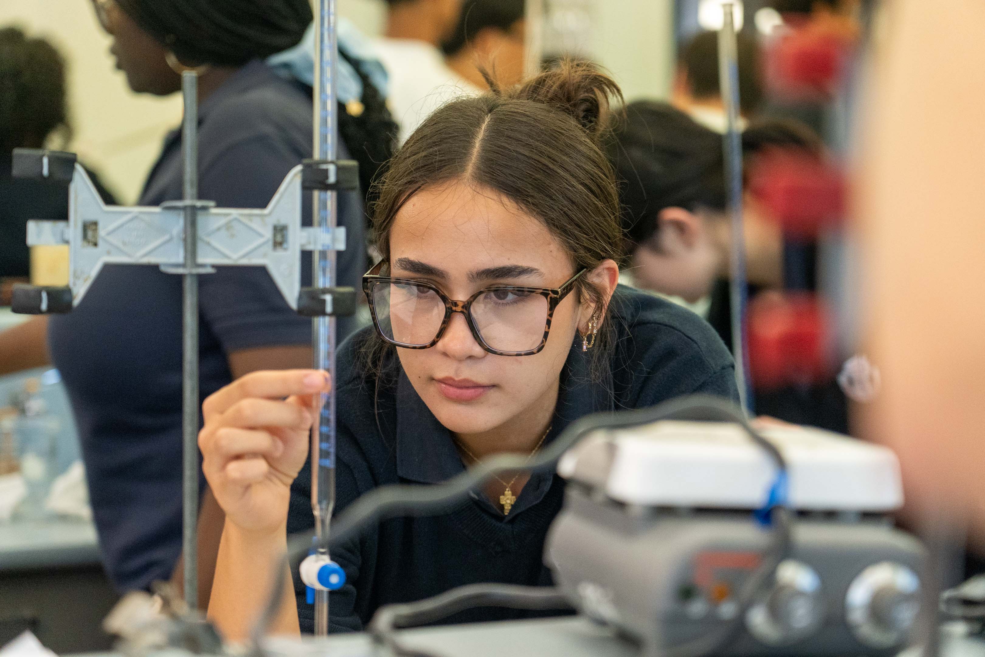 Young woman wearing safety glasses adjusts a laboratory apparatus, focused during a hands-on science experiment in a busy classroom setting.