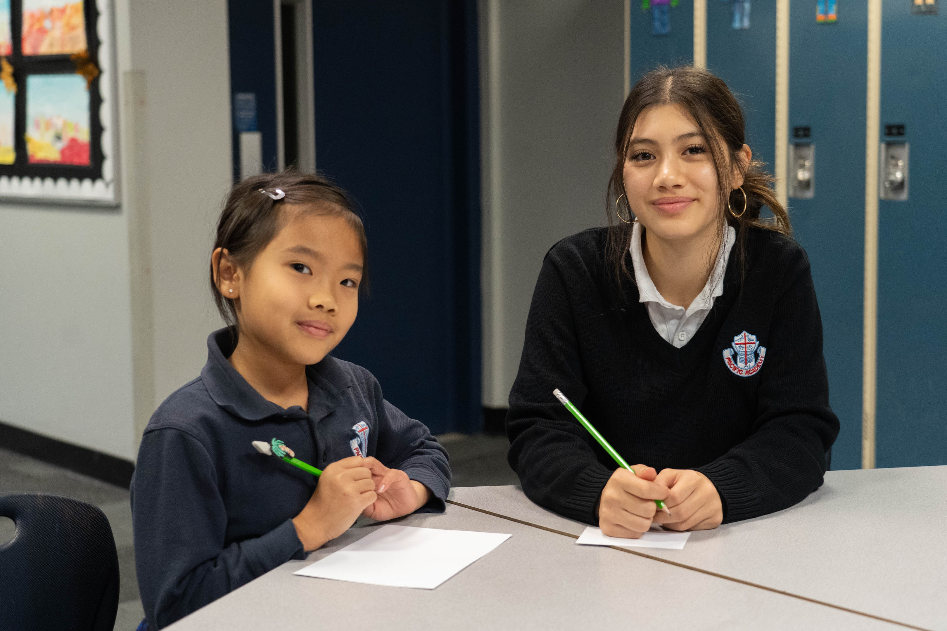 Smiling schoolgirl and teenage tutor sit at a table, holding green pens while writing beside blue lockers in a welcoming classroom.