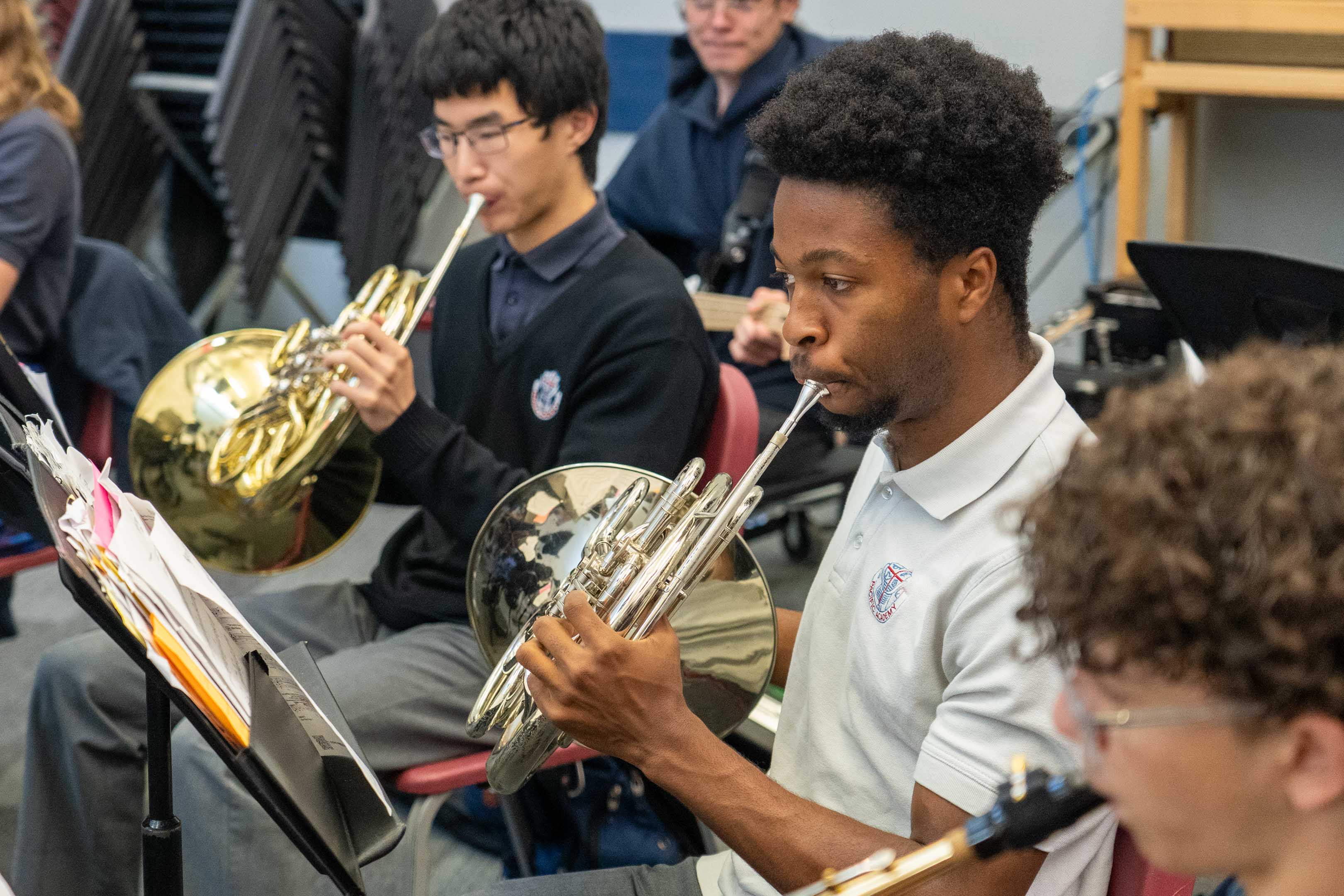 Students play French horns in a school band rehearsal, reading sheet music together in a focused, collaborative atmosphere.