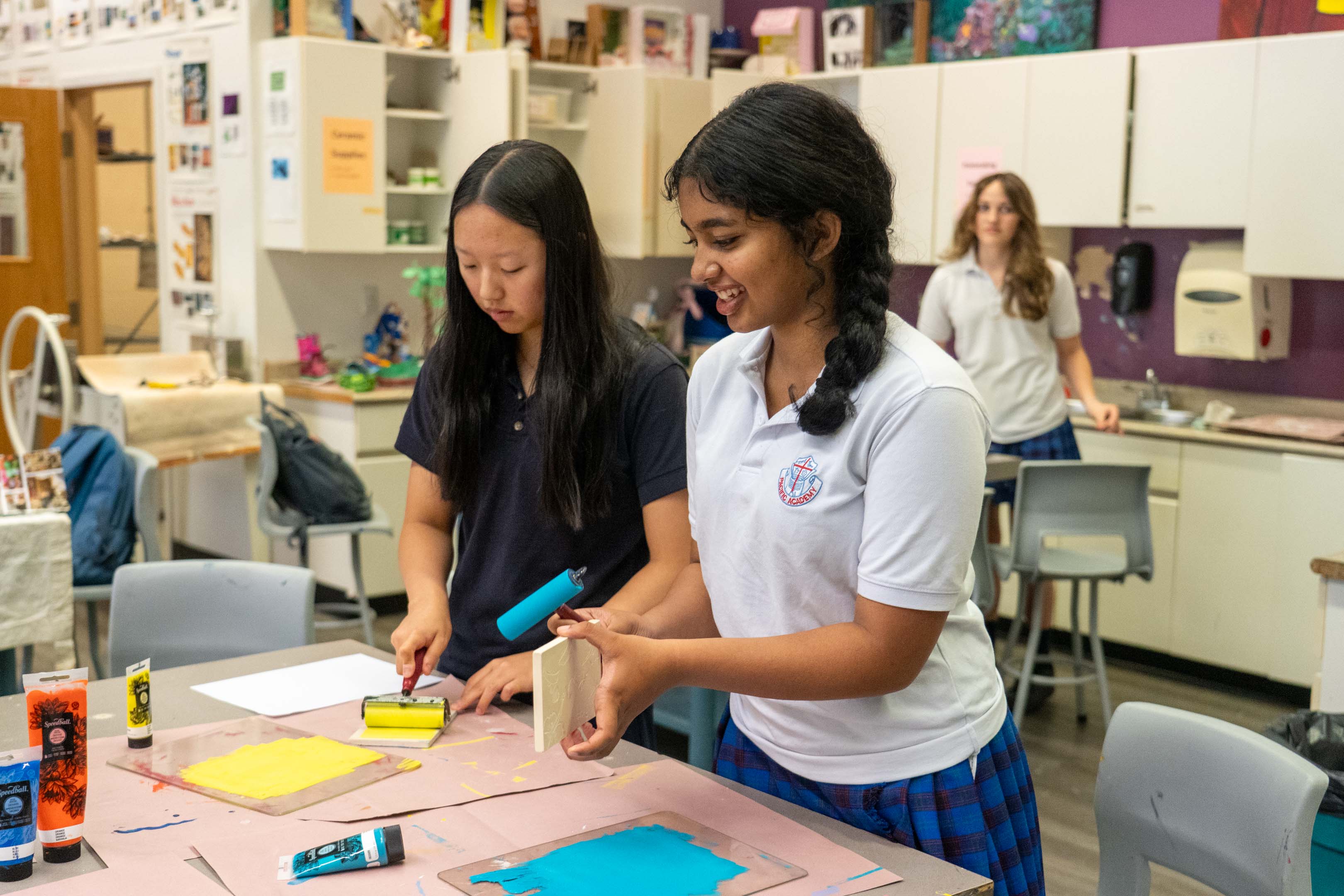 Two students collaborate on a colorful classroom art project, painting paper at a table while classmates and supplies surround them.