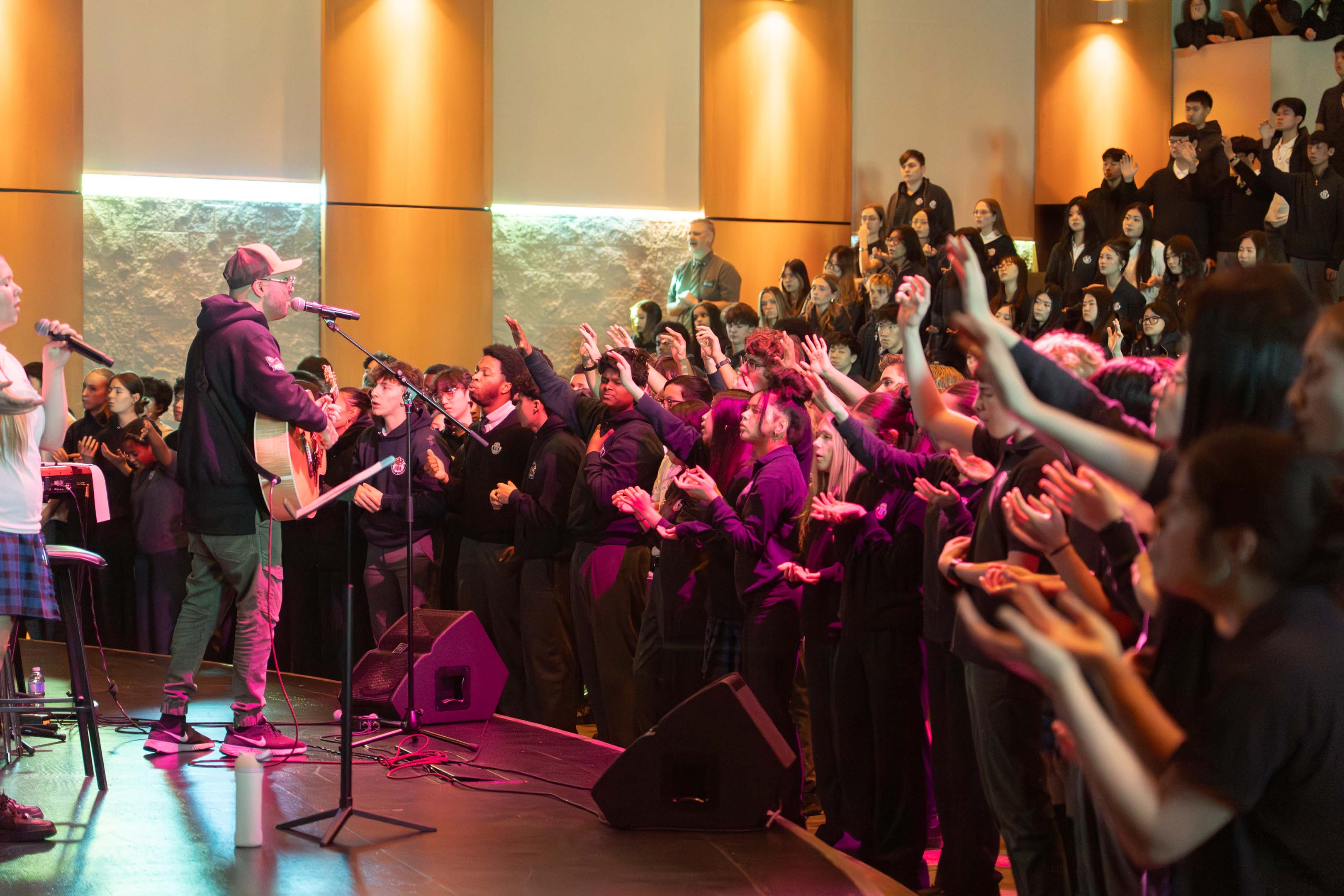 Live concert in a warmly lit venue, where a guitarist sings into a microphone as an enthusiastic crowd reaches toward the stage.