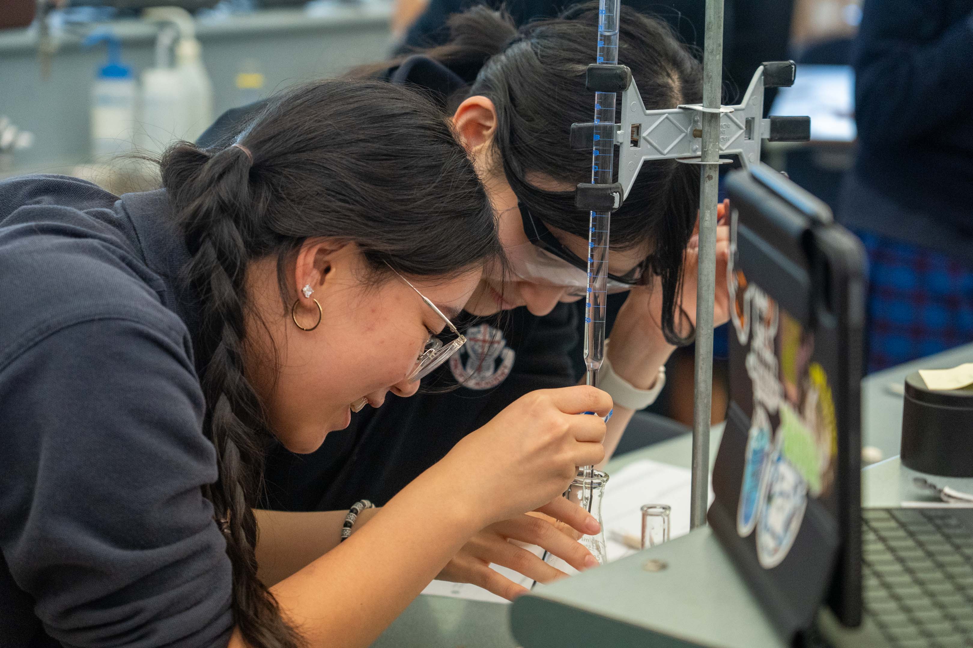 Two students in safety glasses conduct a hands-on science experiment, carefully adjusting lab equipment in a focused classroom setting.