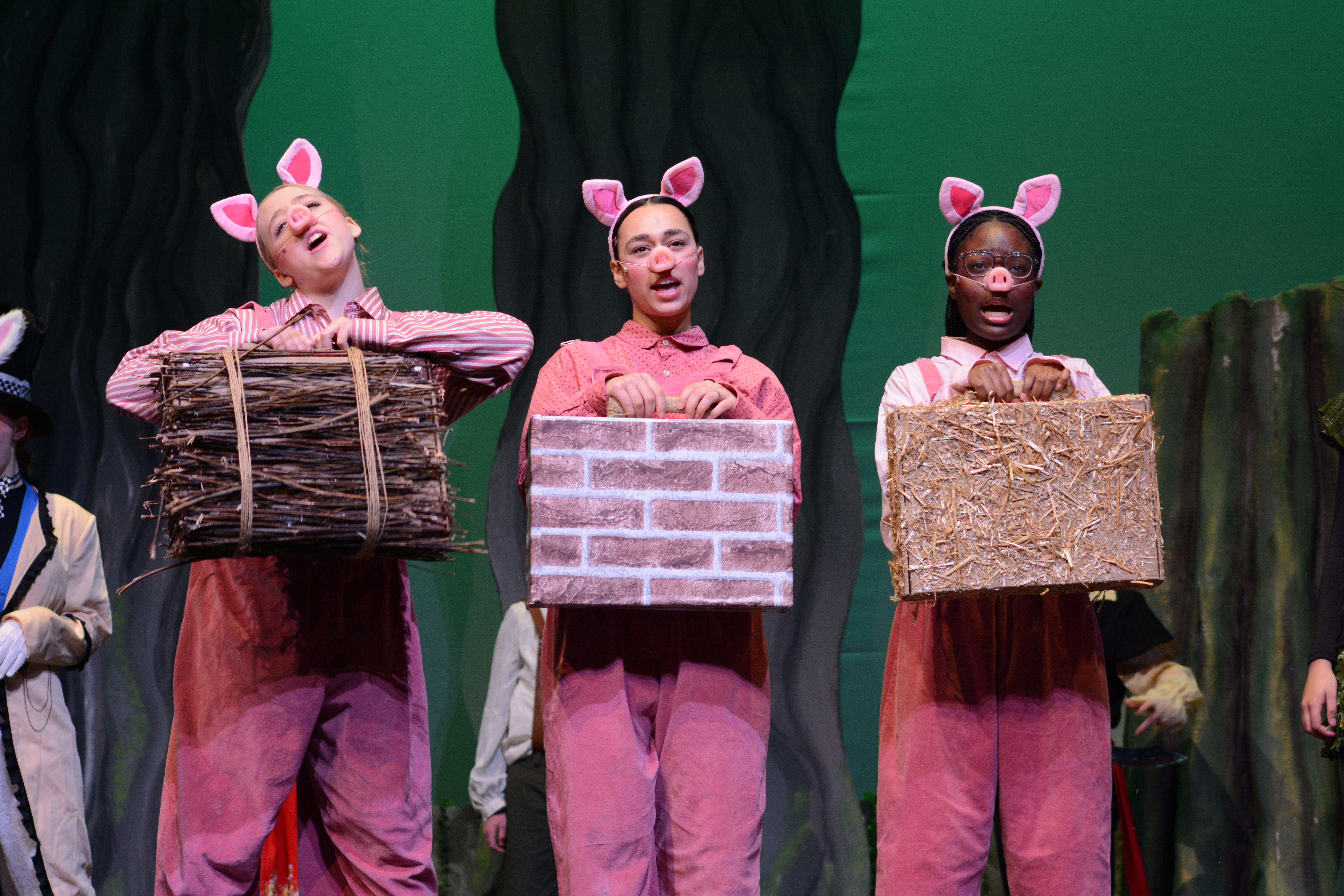 Three performers in pink pig costumes sing onstage, holding straw, brick, and stick house props, creating a playful theatrical scene.
