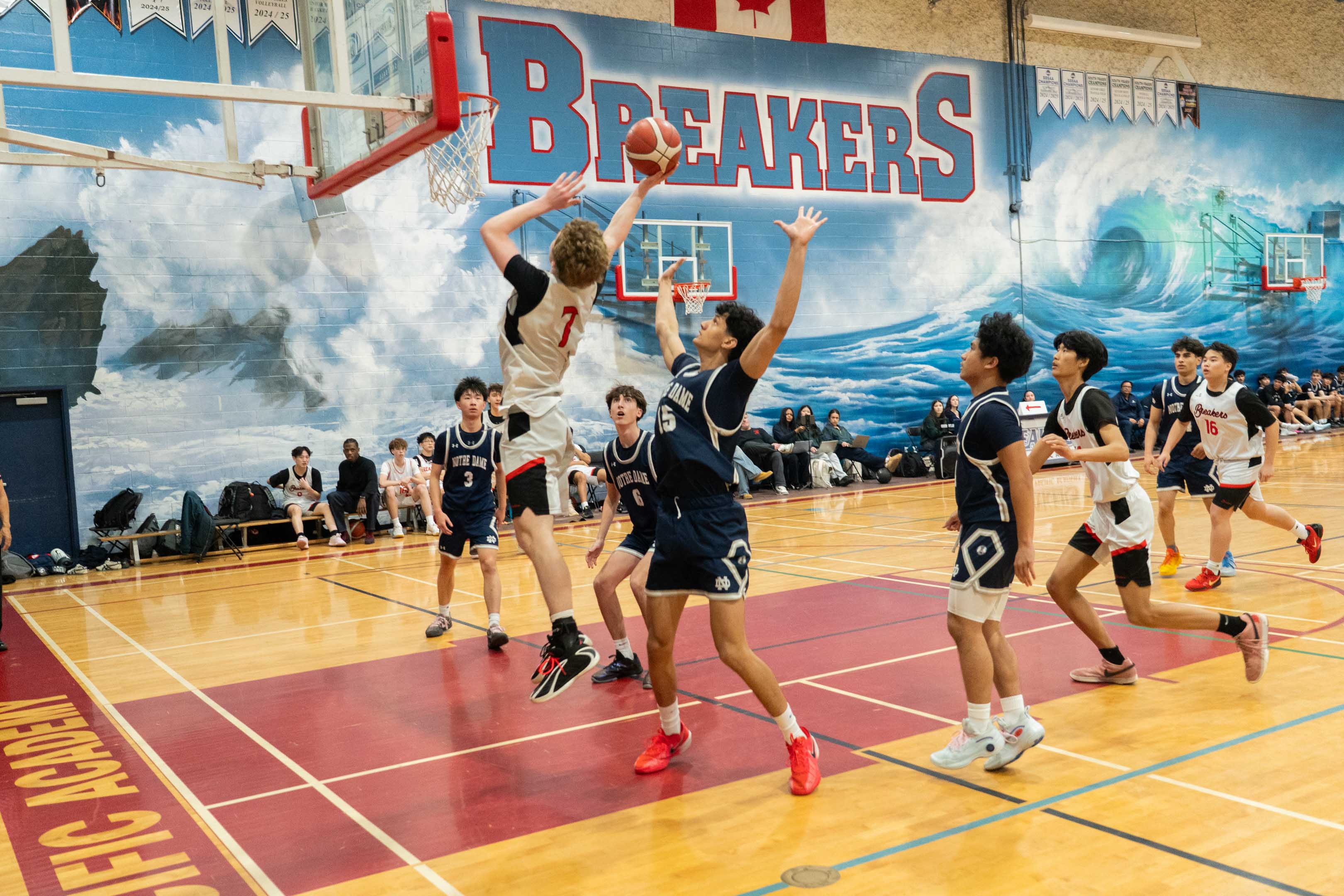 Basketball players leap for a shot beneath the hoop in a lively gym, with teammates defending and “BREAKERS” painted on the wall.