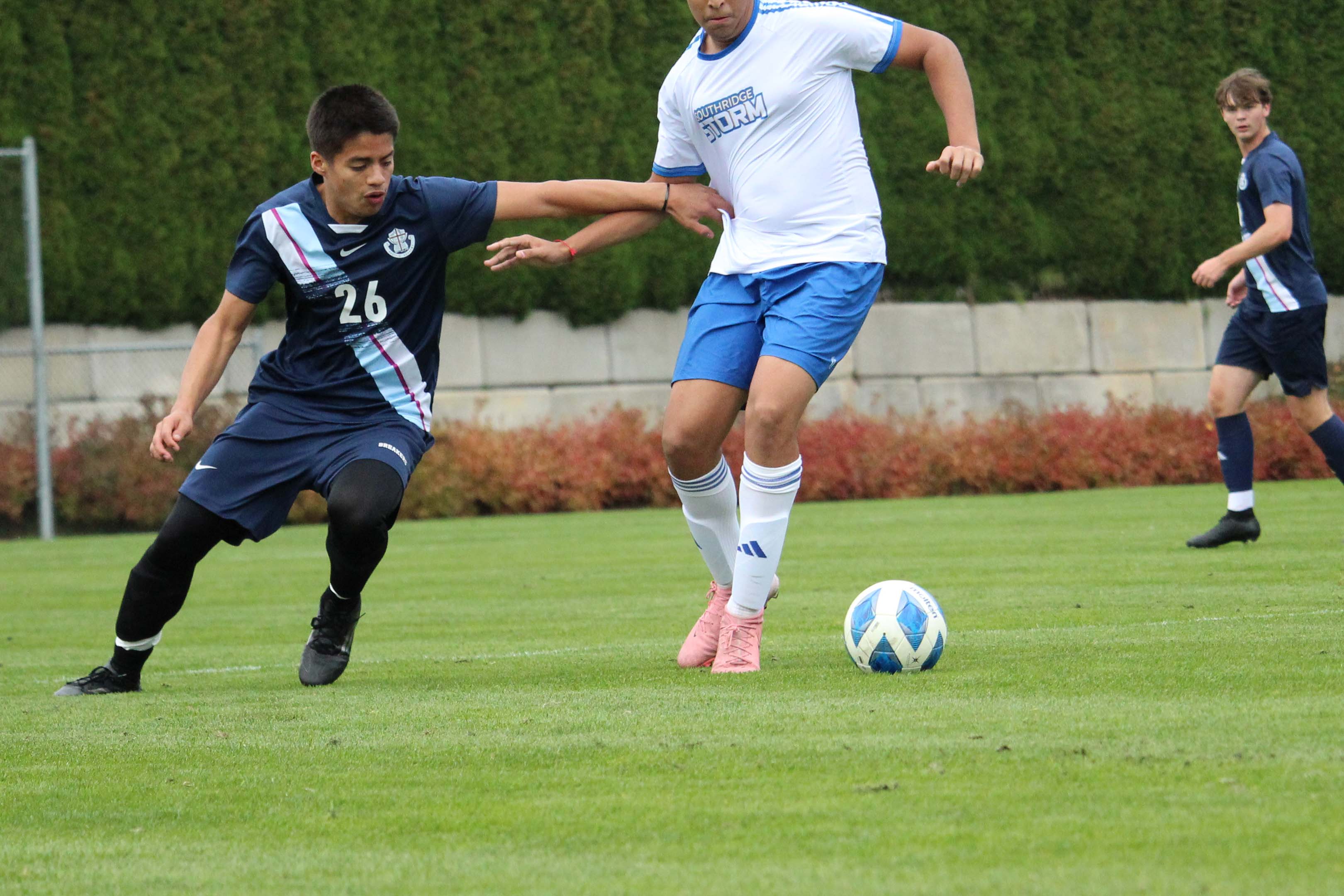 Two youth soccer players battle for the ball on a grassy field; one in navy lunges defensively as another in white and blue dribbles.