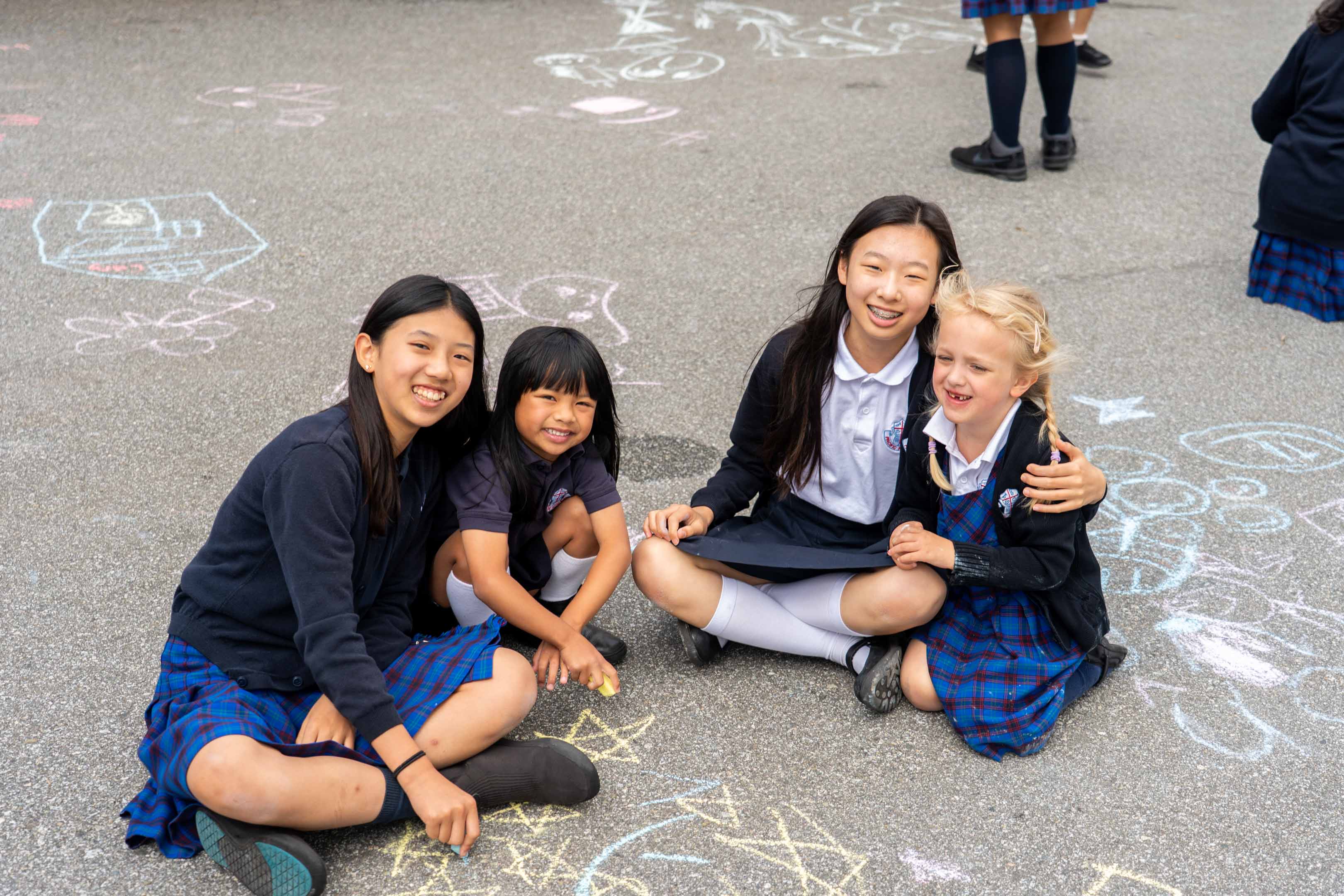 Four smiling schoolgirls in plaid uniforms sit together on a chalk-decorated pavement, posing warmly and joyfully for a photo outdoors.