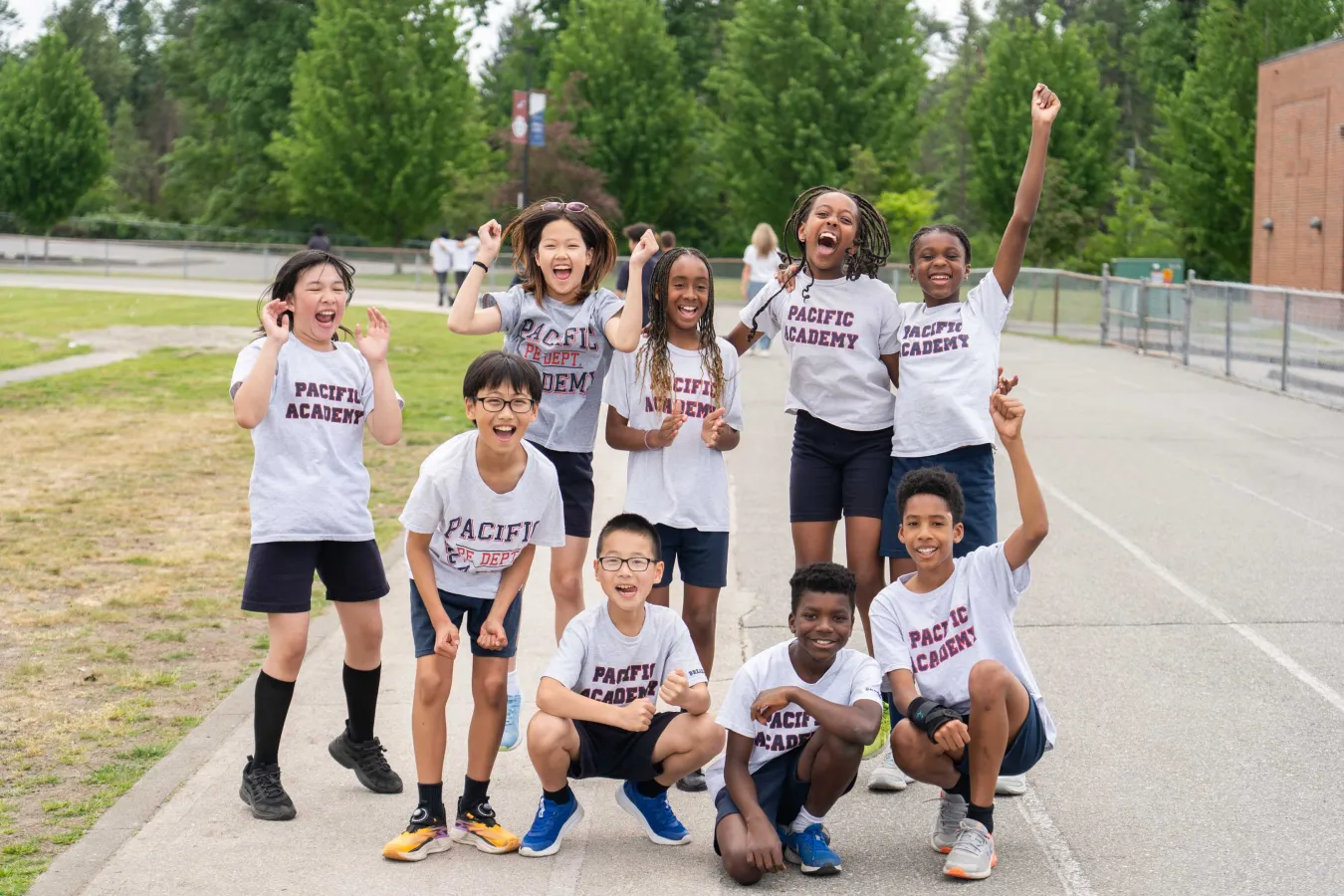 Joyful Pacific Academy students cheer and pose together on a school track, smiling and raising their arms during an outdoor team celebration.