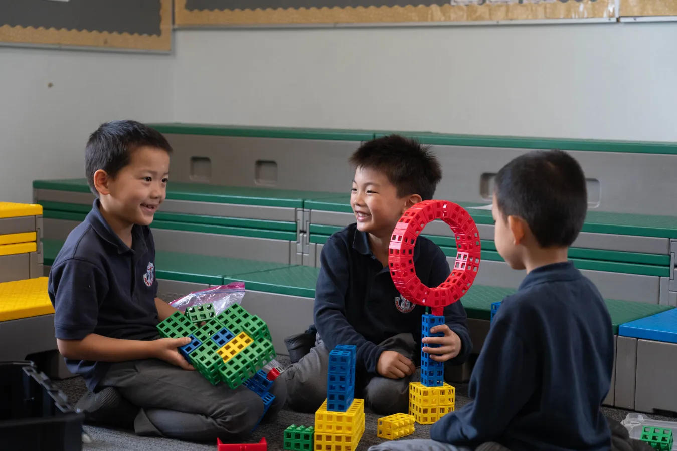 Three schoolboys smile and play with colorful construction toys, building towers and holding a red ring in a bright school classroom.