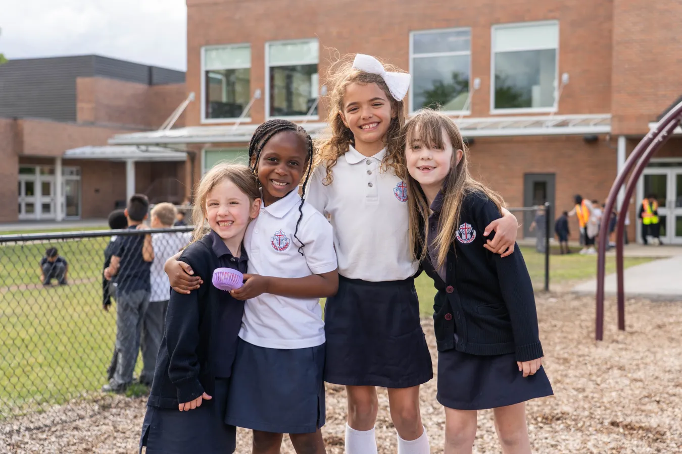 Four smiling schoolgirls in school uniforms stand arm-in-arm outside a brick school, with a playground and classmates behind them.