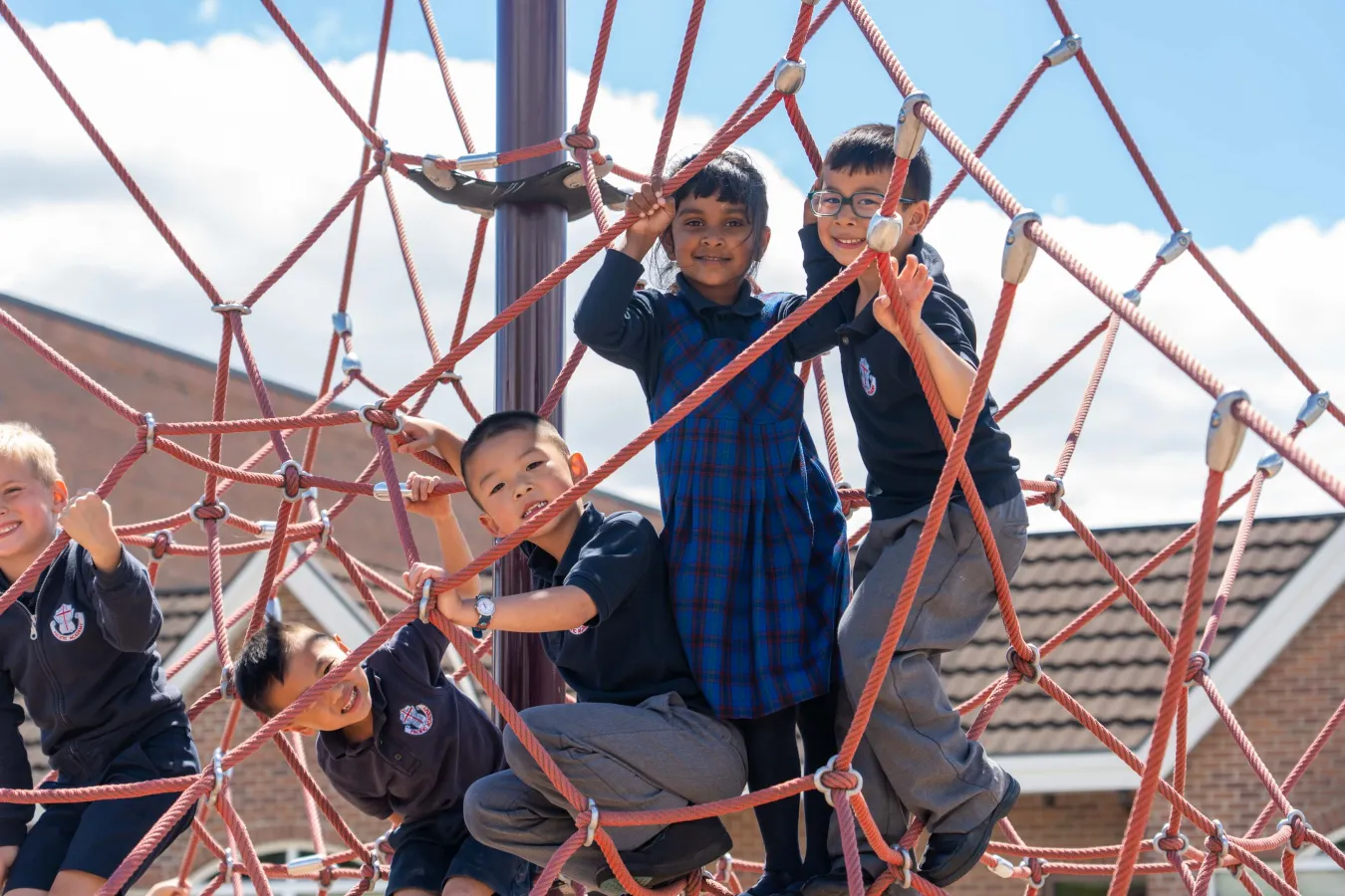 Five smiling schoolchildren climb a red rope playground structure outdoors in school uniforms beneath a bright blue sky, conveying joyful teamwork.