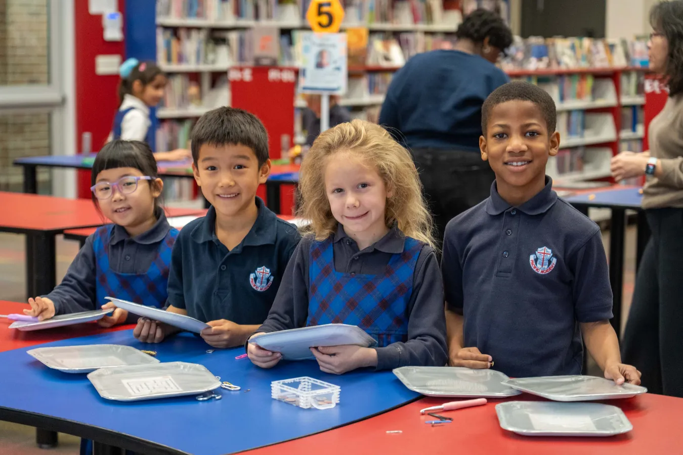 Four diverse elementary students smile at a library table, holding books and papers; colorful shelves and classmates create a cheerful learning scene.