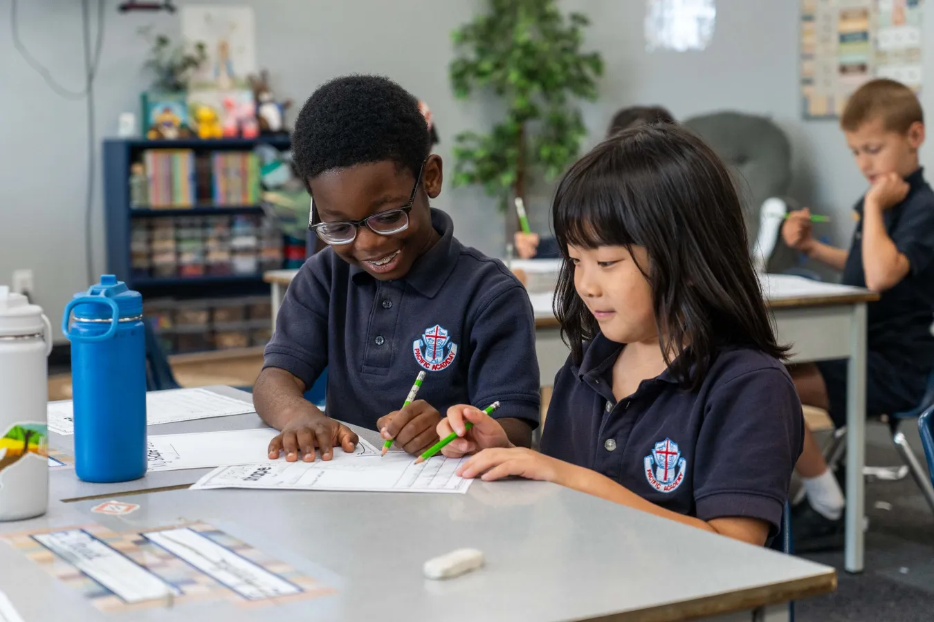 Two elementary students in matching school uniforms collaborate on a worksheet at a classroom table, smiling as they write and color.