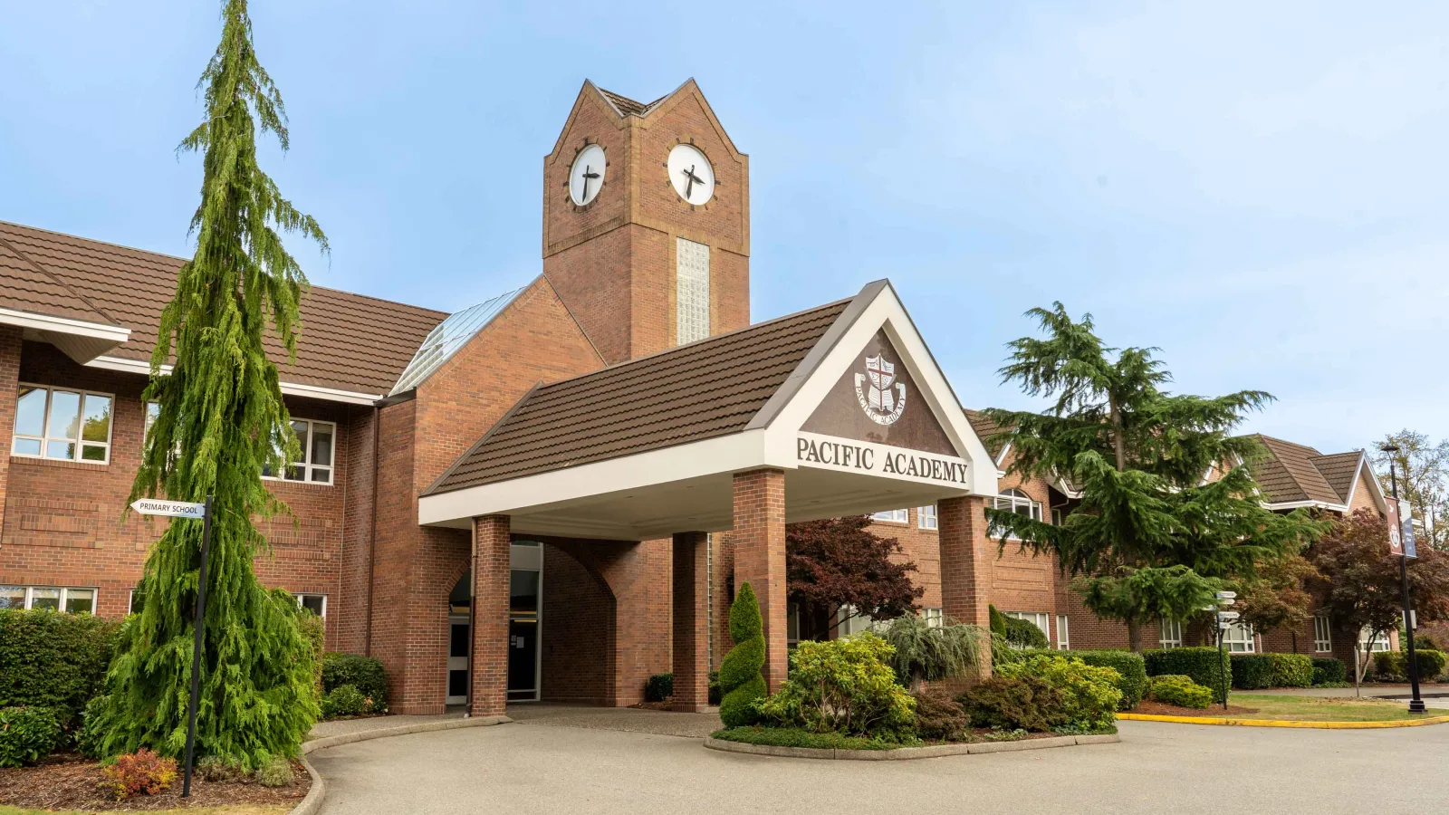 Brick Pacific Academy school entrance with covered driveway, clock tower, trees, and landscaped grounds beneath a bright blue sky.