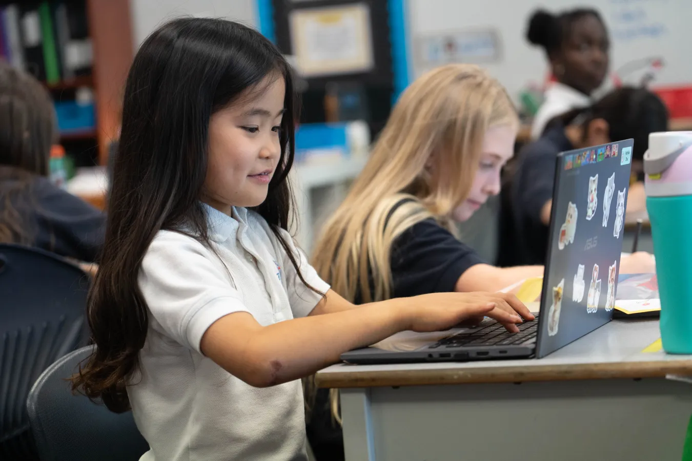 Young students work on laptops in a bright classroom, focused on digital learning while classmates collaborate in the background.