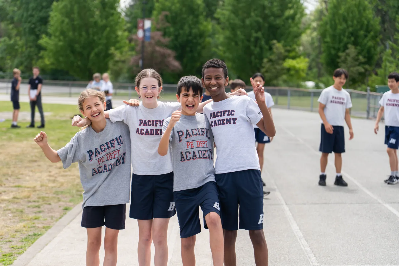 Four cheerful Pacific Academy students pose arm-in-arm on a track, smiling and celebrating; classmates and green trees fill the background.