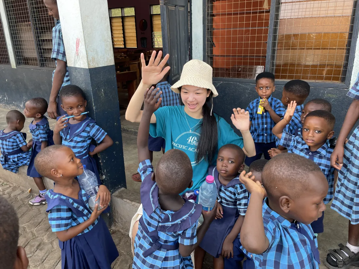 Smiling volunteer in a sunhat waves among children in matching blue plaid school uniforms outside a classroom, creating a joyful scene.