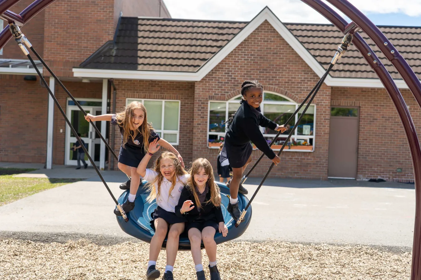 Four children—three girls and one boy—play together on a large basket swing outside a brick school, smiling in bright sunlight.