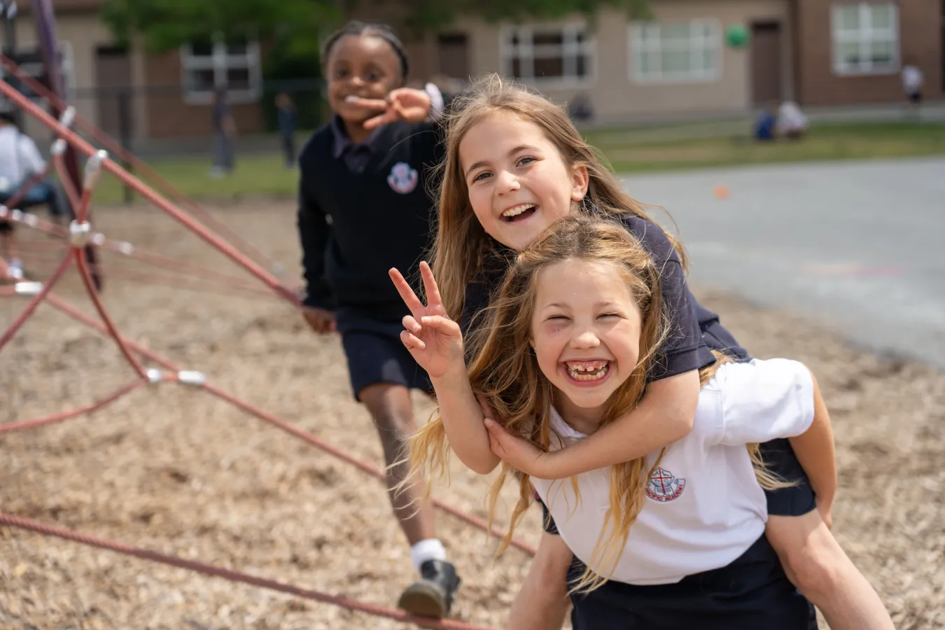 Three joyful schoolgirls play on a playground; one carries another while a third smiles and waves. A school building and climbing frame appear behind them.