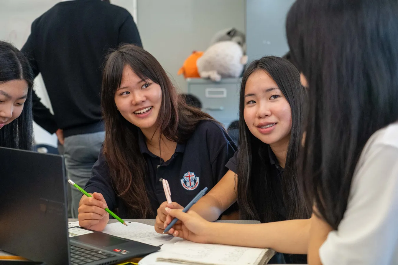 Three smiling students collaborate around a laptop and worksheets in a classroom, writing and discussing ideas in a friendly, focused atmosphere.