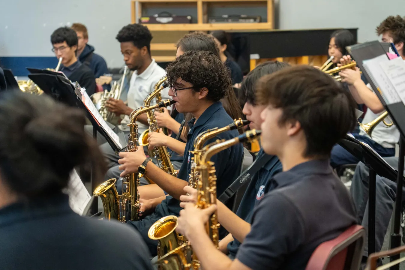 Student musicians rehearse saxophones in a school band, reading sheet music together in a focused, collaborative classroom setting.