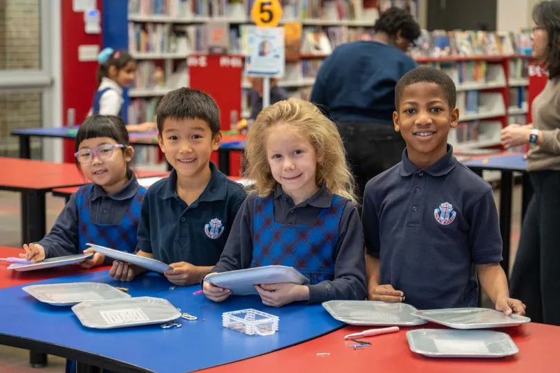 Four diverse elementary students smile at a library table, holding books and papers; colorful shelves and classmates create a cheerful learning scene.