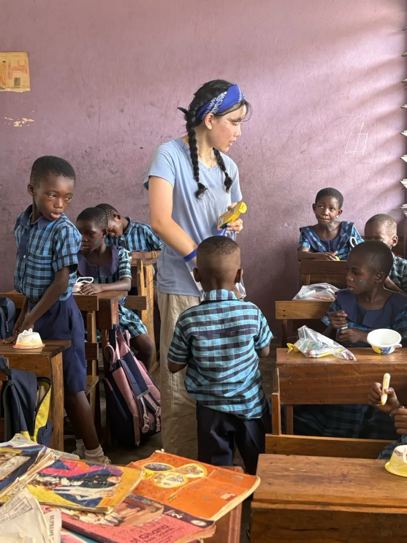 A woman in a blue headscarf helps Black schoolchildren in a warm classroom, surrounded by wooden desks, books, snacks, and backpacks.