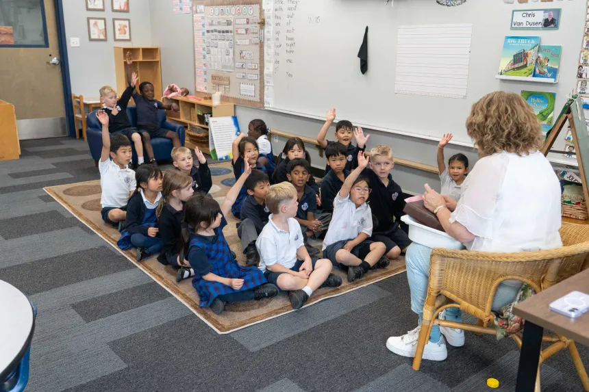 Teacher reads to a diverse group of young students seated on a classroom rug; many raise their hands, creating an engaged, cheerful scene.