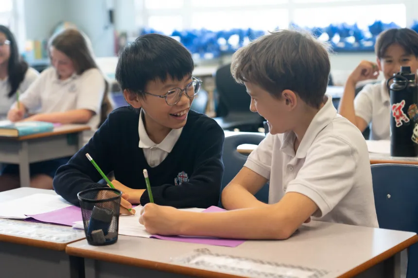 Two smiling schoolboys collaborate at a classroom desk, writing on colorful papers; classmates and a water bottle appear behind them.
