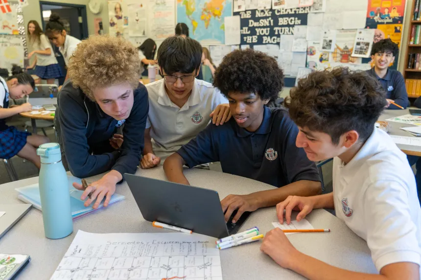 Four diverse students collaborate around a laptop and worksheets in a classroom, focused on a group project with an engaged, friendly tone.