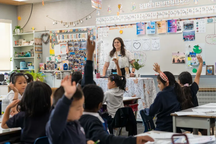 Teacher leads an engaging classroom discussion as diverse elementary students raise their hands, surrounded by colorful learning displays and artwork.