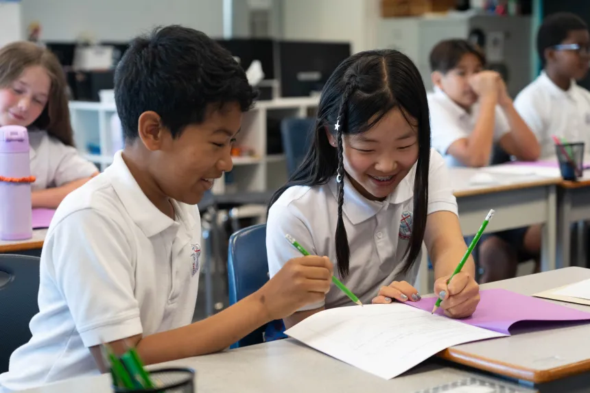 Two smiling students collaborate on a classroom worksheet, writing with colored pencils at a desk while classmates work in the background.