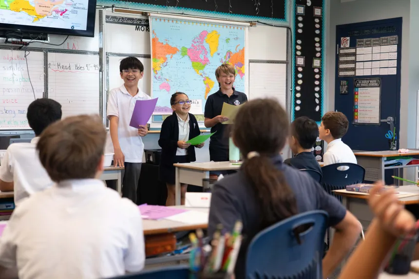 Students present in a lively classroom, holding colored papers before a world map while classmates watch, creating an engaged, cheerful mood.