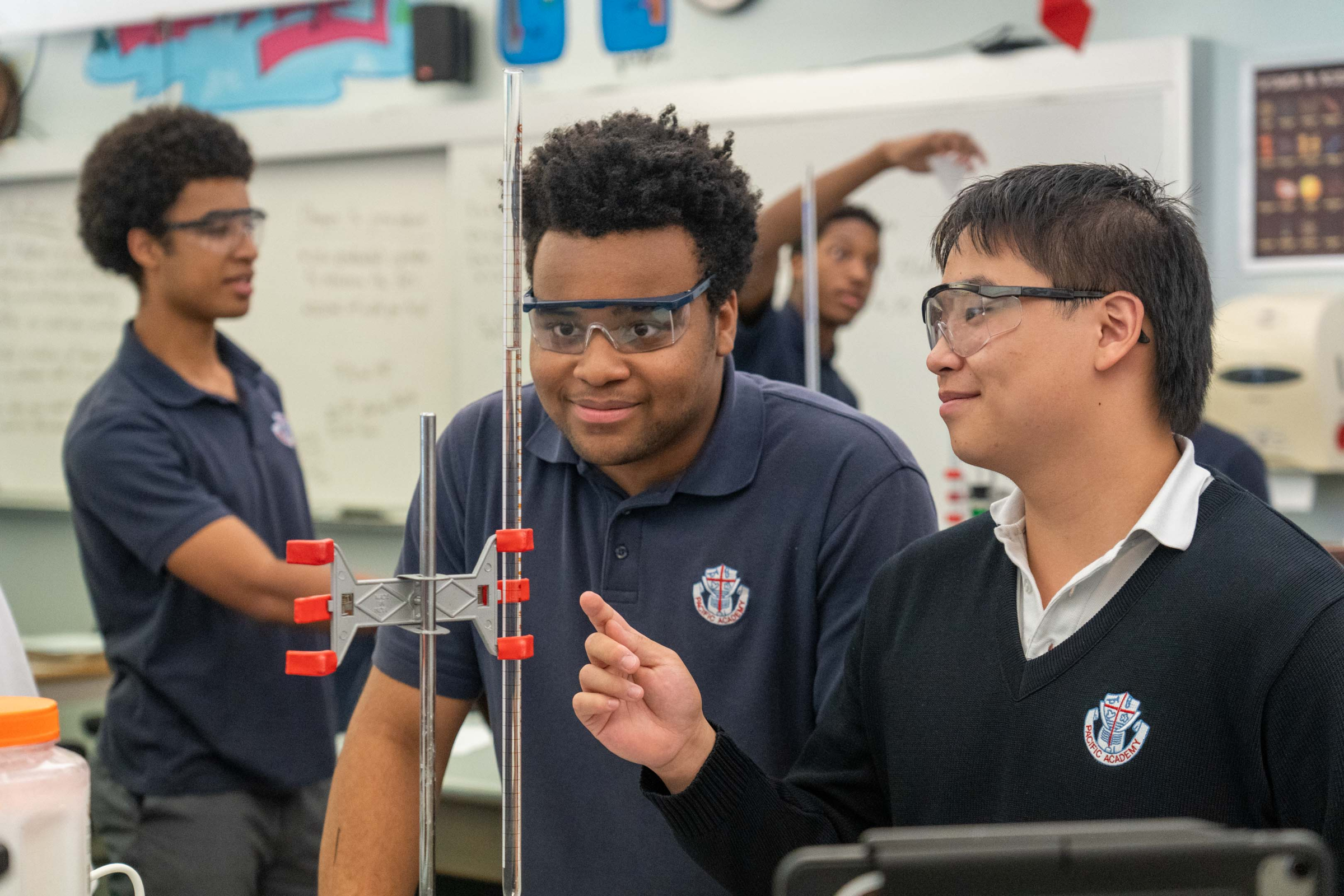Students in safety goggles collaborate around a lab apparatus, smiling and focused in a classroom science experiment.