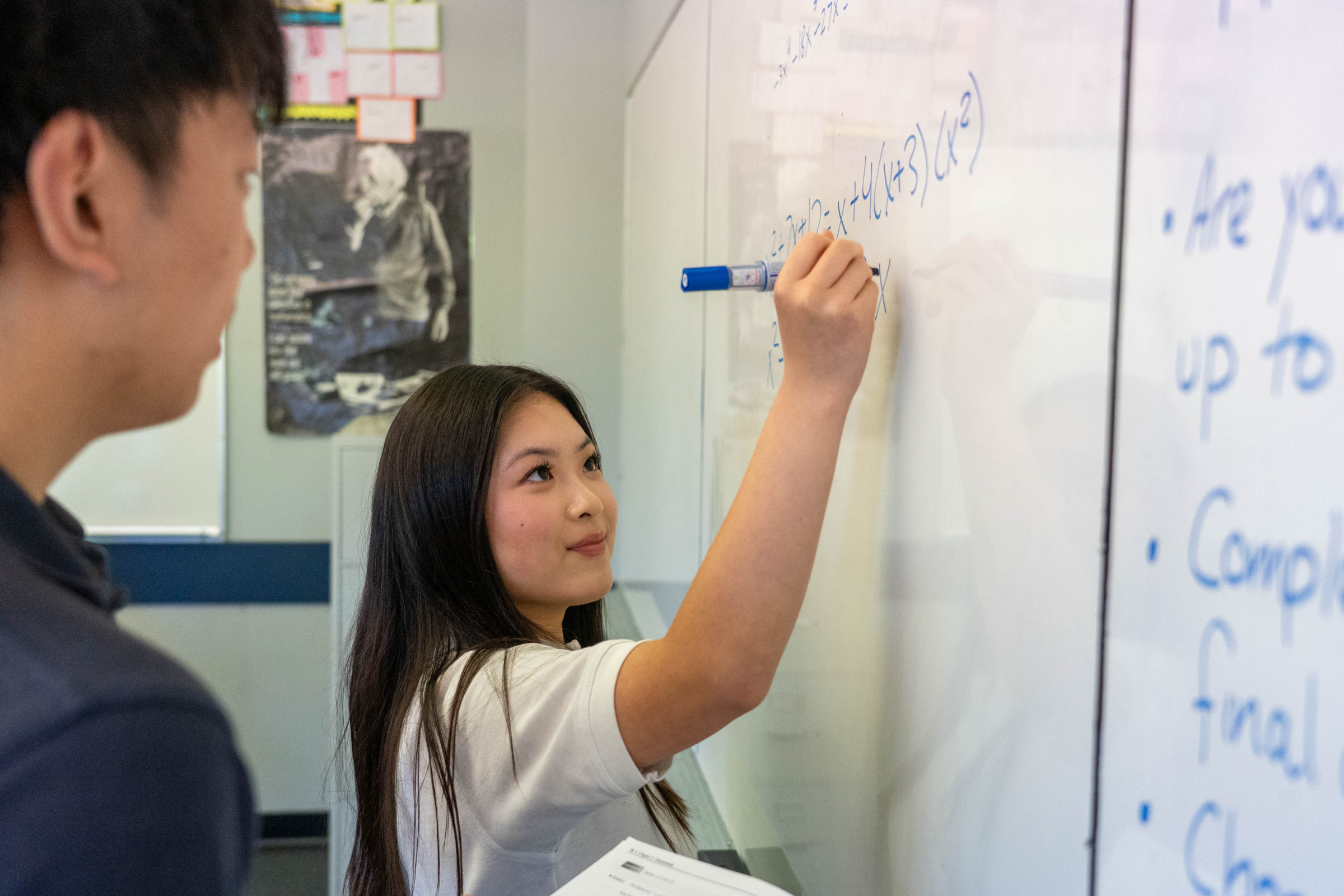 Young woman writes notes on a whiteboard with a blue marker as a colleague watches, conveying a focused, collaborative meeting.
